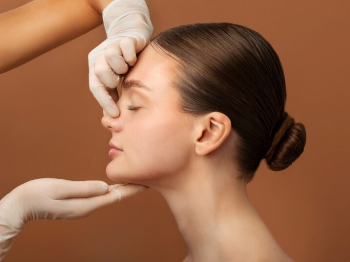 Surgeon touching woman nose, consulting lady before rhinoplasty operation, brown studio background, side view. Plastic surgery concept