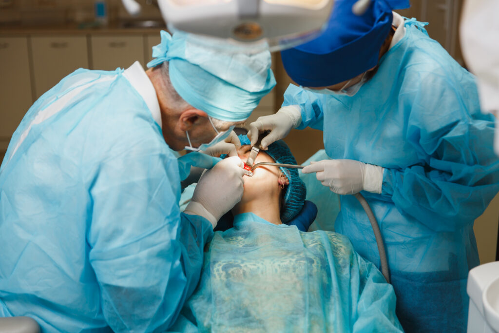 Male old professional dentist surgeon in uniform and female assistant helps to performing operation install dental implant teeth of woman patient in clinic light office with modern tools equipment.