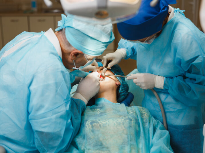 Male old professional dentist surgeon in uniform and female assistant helps to performing operation install dental implant teeth of woman patient in clinic light office with modern tools equipment.